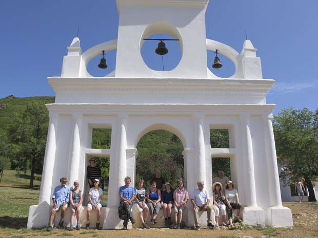 A white facade with three arches and two bells frames the backdrop for a group of people seated or standing, surrounded by greenery under a clear blue sky.